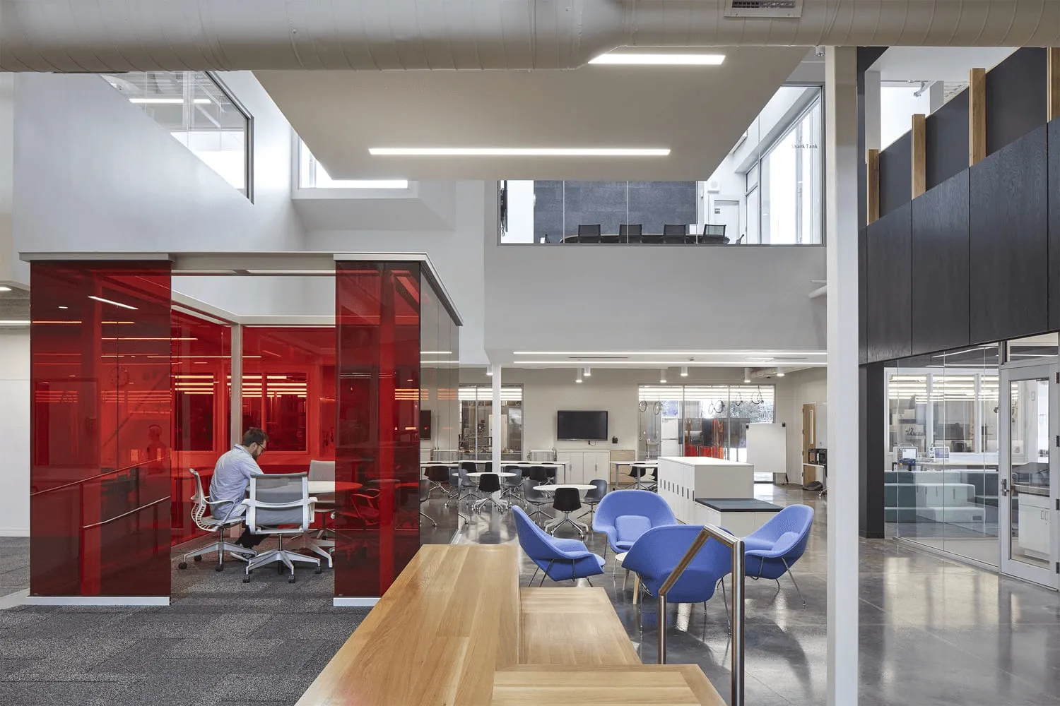 Open office atrium with red glass meeting rooms, blue lounge chairs, and exposed ductwork.