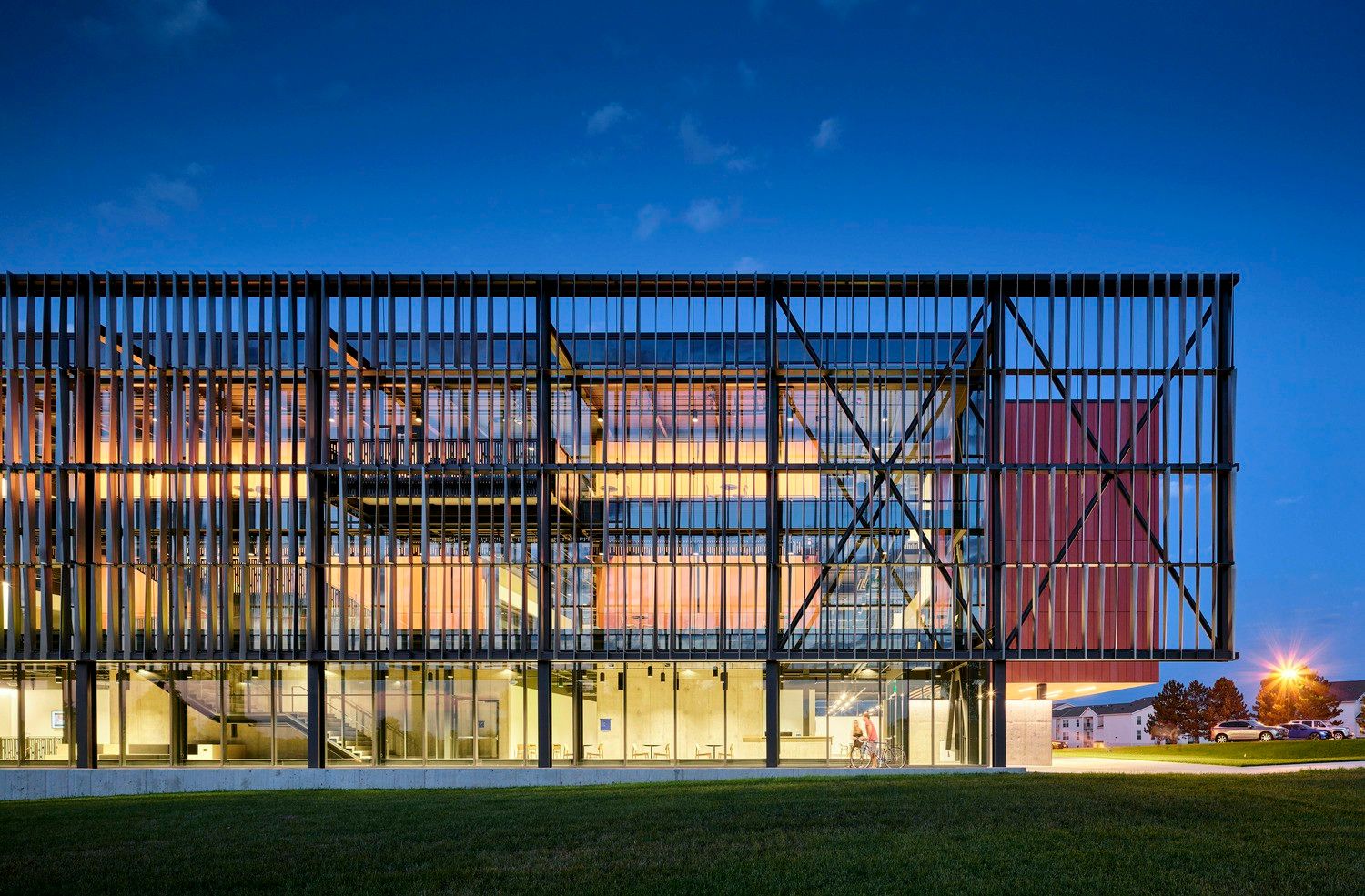Modern three-story academic building at dusk with illuminated interior visible through floor-to-ceiling glass walls. The exterior features black structural beams in a grid pattern and a red accent wall. A grassy lawn extends from the building's base.