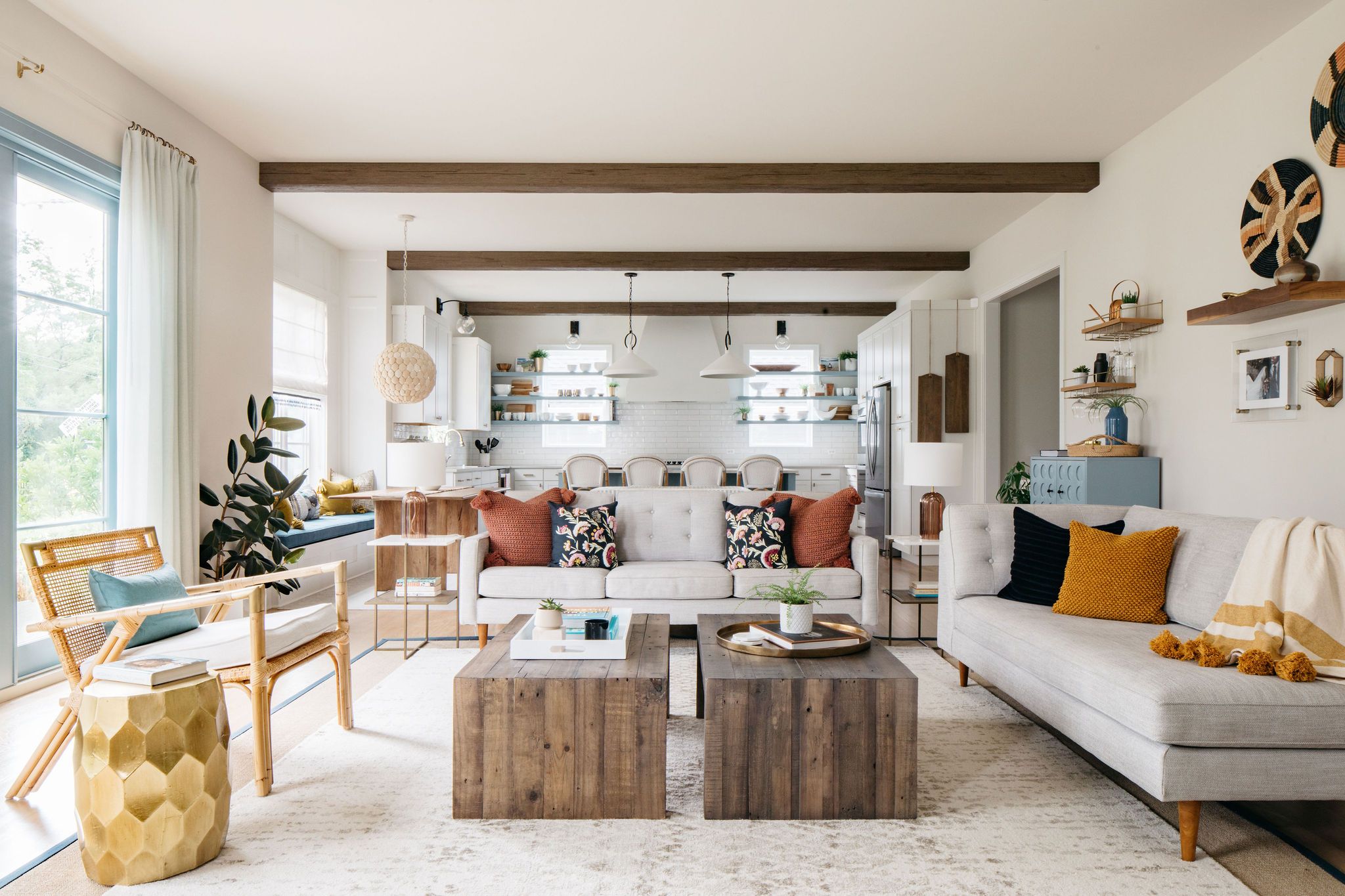 Open-concept living space and kitchen designed by Outline Interiors featuring a modern bohemian aesthetic. The room includes exposed wooden beams, light gray sofas with colorful accent pillows, rustic wooden coffee tables, and a rattan accent chair. The white kitchen in the background has open shelving, while decorative elements include a woven pendant light, potted plants, and wall-mounted baskets.