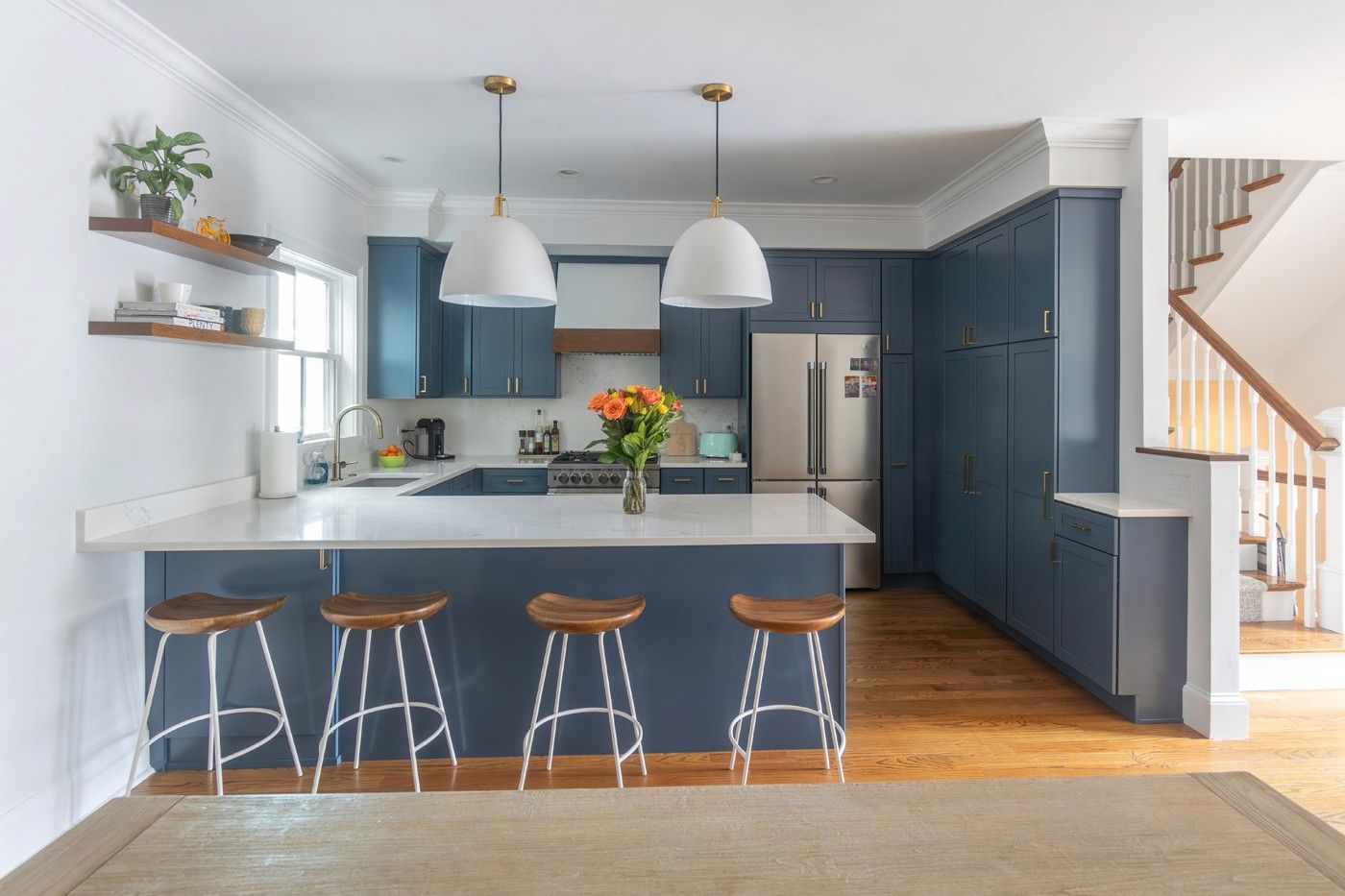 Modern kitchen renovation featuring slate blue cabinets, white quartz countertops, and a peninsula with four wooden barstools. Large white dome pendant lights hang above, and the space includes floating wooden shelves, stainless steel appliances, and hardwood floors. A staircase with wooden treads is visible to the right.