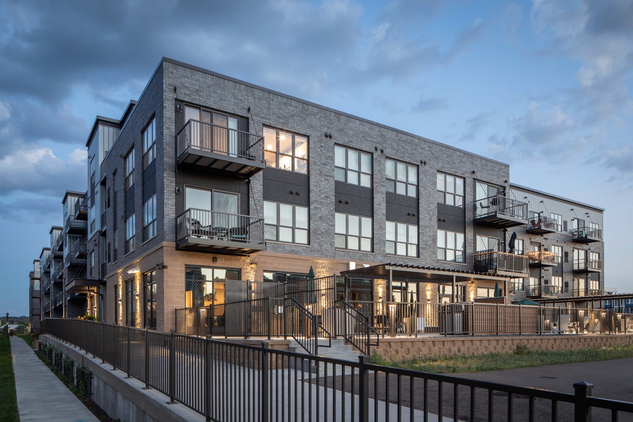 Modern apartment complex photographed at dusk featuring a mix of grey brick and dark metal panels. The four-story building showcases private balconies, large windows, and ground-floor patios with decorative uplighting along the columns. An elevated walkway with black metal railings runs along the front of the building.
