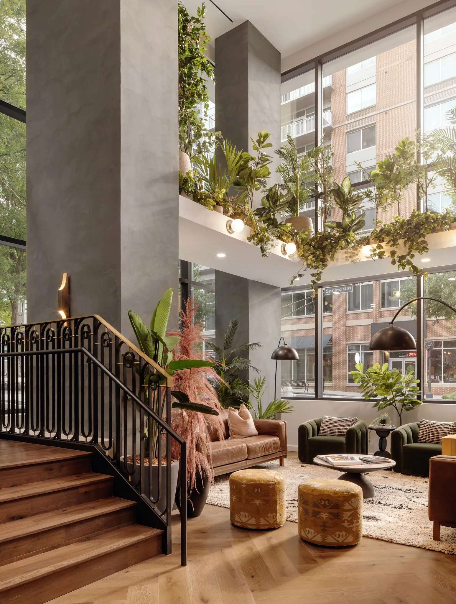 Double-height lobby seating area with a staircase, tall windows, and dense indoor planting.